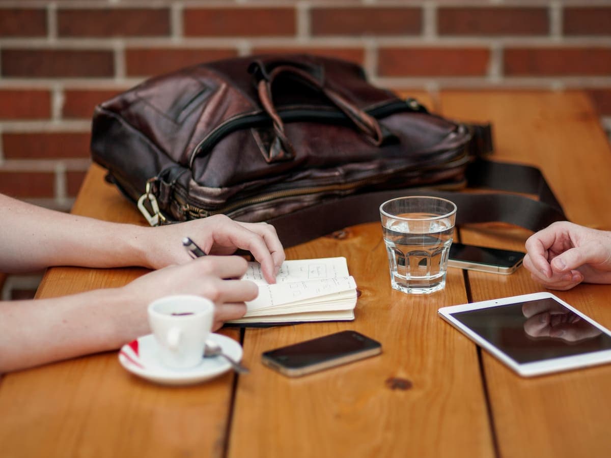 Dua orang berdiskusi di meja kayu dengan buku catatan, tablet, ponsel, dan secangkir kopi