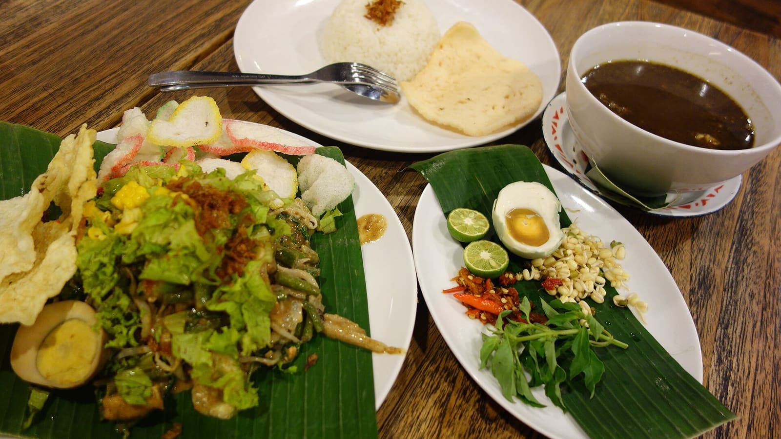 Meja kayu penuh hidangan: pecel beralas daun pisang, semangkuk rawon, nasi putih, dan lalapan